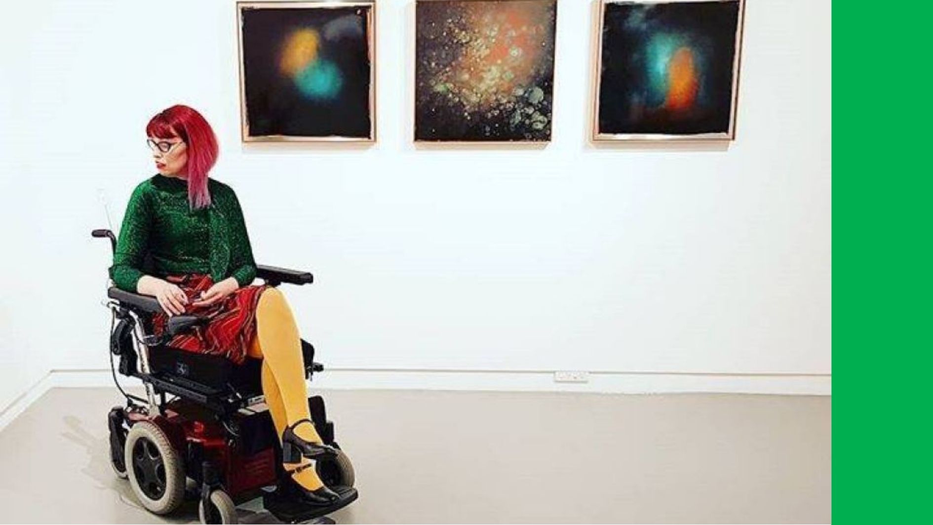 Image Description: A wheelchair user with pink hair and bright clothing is at an exhibition in front of three colourful artworks.